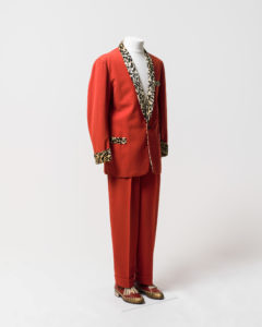 Mens suit consisting of jacket and trousers, worn by Johnny O'Keefe. -1 Jacket made of red fabric with black and cream coloured 'leopard print' velvet trimming at collar, cuffs and pockets. The jacket has a shawl collar with the collar and lapels lined with leopard print velvet which extends on the inside of the jacket to the waist. The jacket fastens at the waist with a single black plastic button which is sewn to the jacket with red cotton. The jacket is long sleeved and the cuffs are trimmed with leopard print velvet. The jacket has a fake external left breast pocket and two fake external waist pockets on the left and right sides, all pockets are trimmed with leopard print velvet. There are no internal pockets. The inside of the jacket is lined with a red [silk] fabric and leopard print velvet lines the collar and lapel area extending to the waist. The shoulders are slightly padded. The jacket has several moth holes including two on the back of the right shoulder and two below the button. The lining has a hole at the neck and is torn at each armhole and has detatched from the velvet below the buttonhole. The velvet lining is worn around the neck and above the button. -2 Trousers made of red fabric, matching trousers to jacket (98/32/2-1). Trousers have 2 side pockets and a coin pocket at the waist on the right side. Inside of pockets are made of a cream cotton fabric. Trousers are pleated at the waist at the front and back and have 4 wide belt loops formed in the pleats and one thin belt loop and the centre back of the waist which has a safety pin attached. Trousers fasten at the centre front with a metal zipper, a metal hook and eye and an internal button on the left side of the waist which fastens to a buttonhole on a triangular extension of red fabric on the right side. The trousers are unlined except for the inside waistband which is lined with a band of cream ribbed silk and the zipper which is lined with black [silk] fabric. The trousers are hemmed to give the appearence of turned up cuffs at the hem of each leg. The fabric forming the inside of the right pocket has a handwritten inscription in black ink, 'O'Keefe's [sic]/3483'. There are white marks on the legs and repairs have been made to the crotch area.